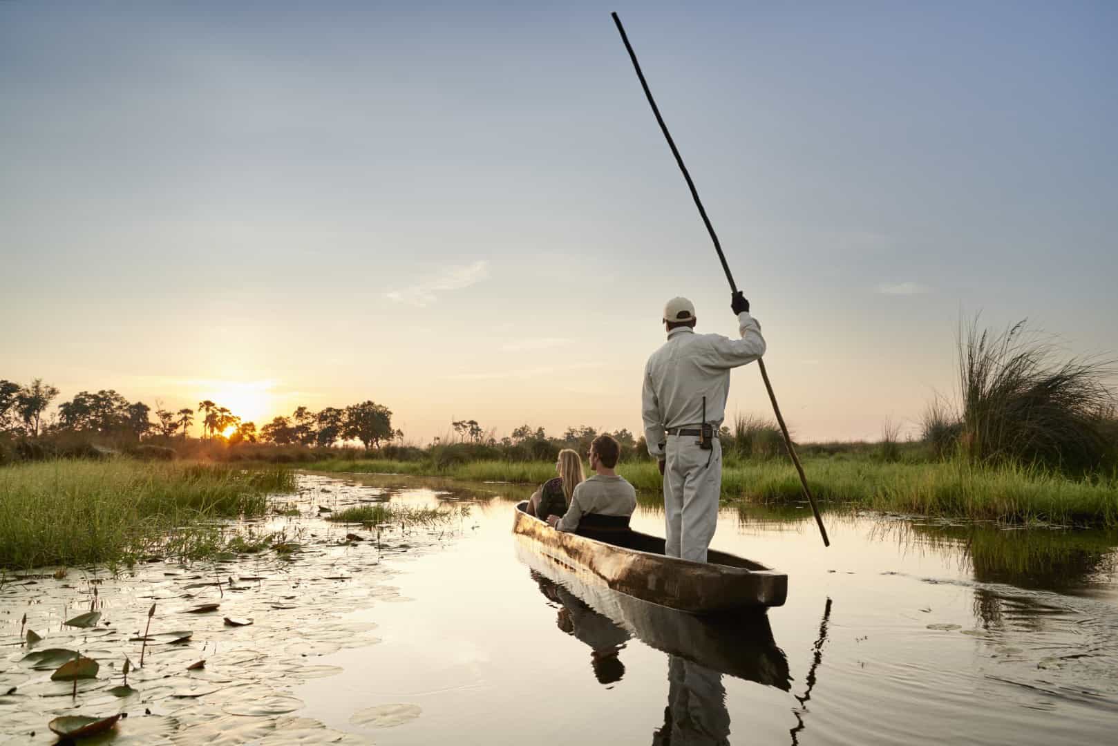 A man and woman on a mokoro safari in the Okavango Delta with a safari guide, as the sun sets. 