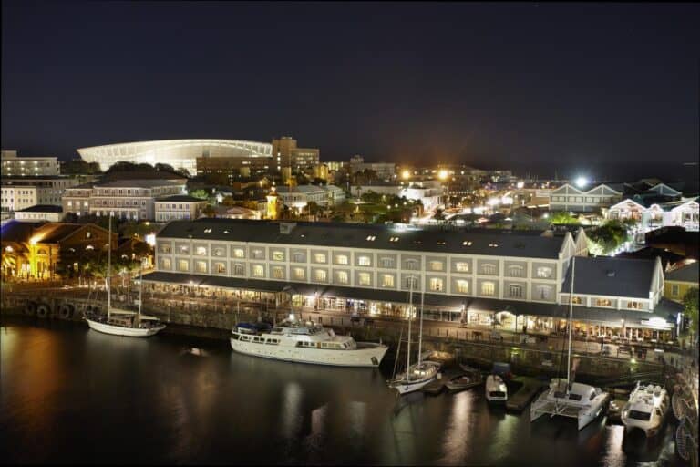 View of Victoria & Alfred Hotel in South Africa at night, featuring boats in the dock.