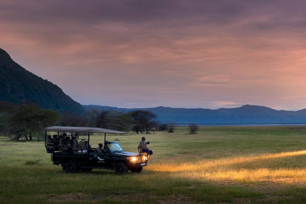 Guests on a dusk game drive in an enclosed safari vehicle with its headlights illuminated as the sun sets over the horizon near andBeyond Lake Manyara Tree Lodge, Tanzania.