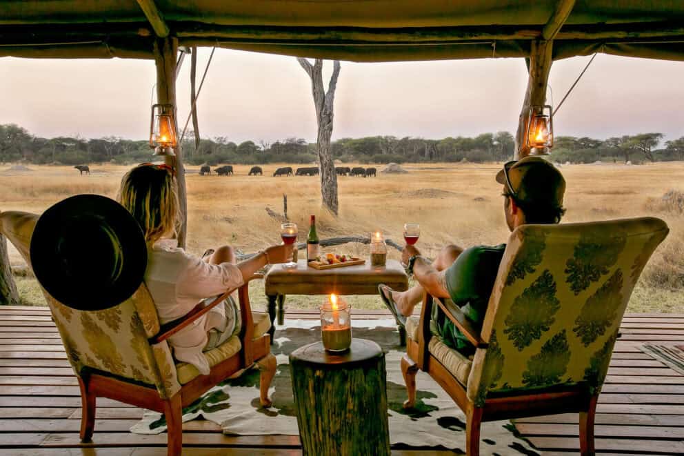 A couple relaxing in comfy cushioned wooden armchairs on a wooden deck watching a herd of buffalo in the distance at The Hide, Zimbabwe.