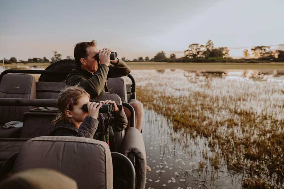 A father and young daughter in a safari vehicle observing the wildlife through binoculars at Vumbura Plains, Botswana.