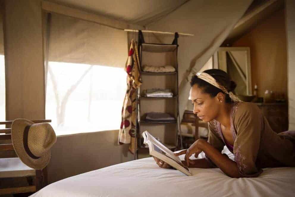 A woman laying on a bed reading a book in a traditional safari tent at Serian's Serengeti Lamai, Tanzania.