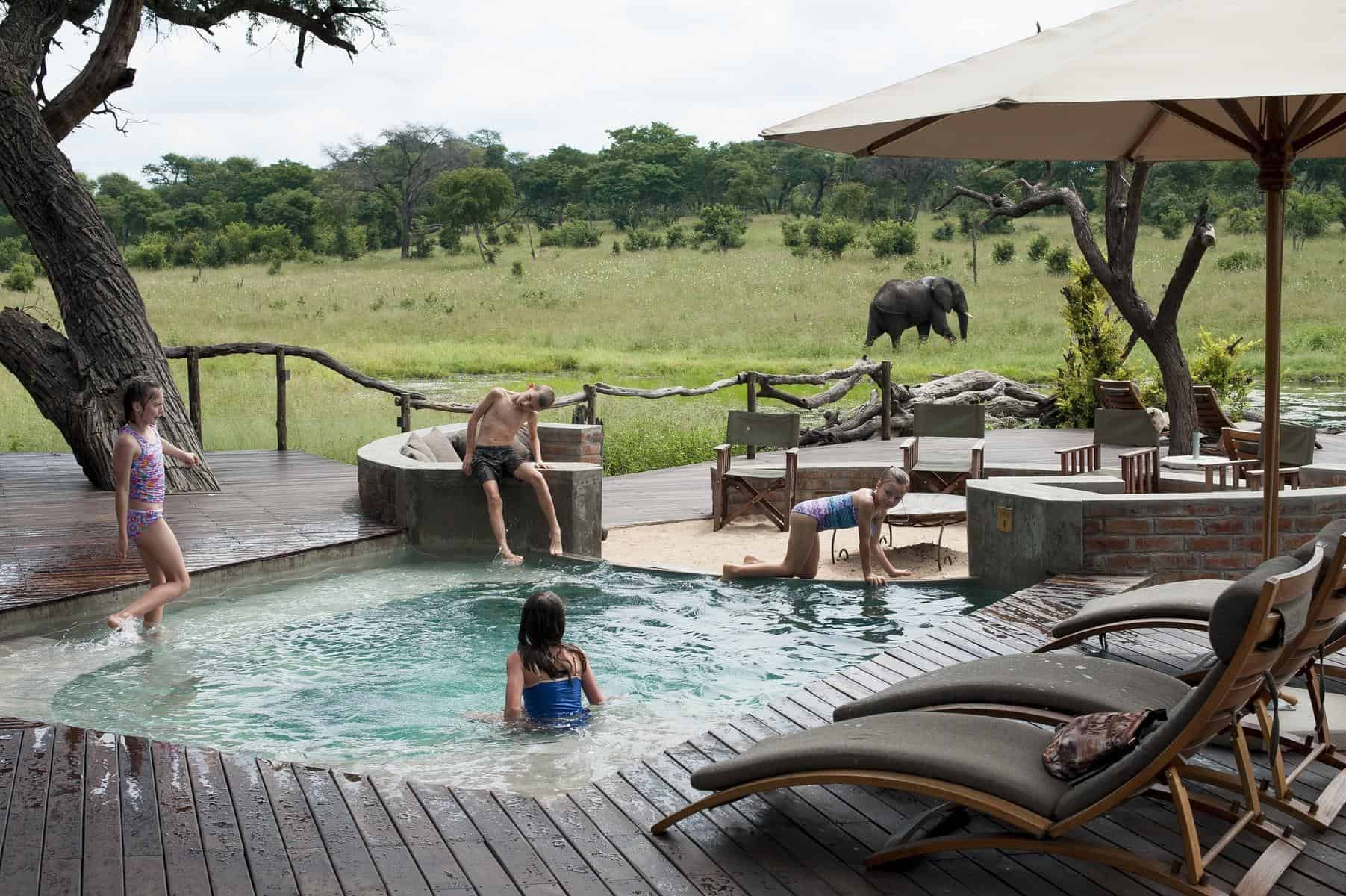 Children playing in a pool with an elephant roaming in the distance at Somalisa Acacia Camp
