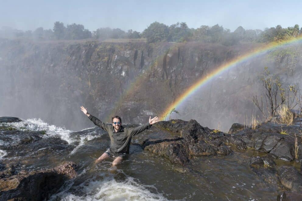 A tourist sitting on the rocky ledge of Devil's Pool, at the edge of Victoria Falls with a rainbow in the back at Tongabezi Lodge, Zambia.