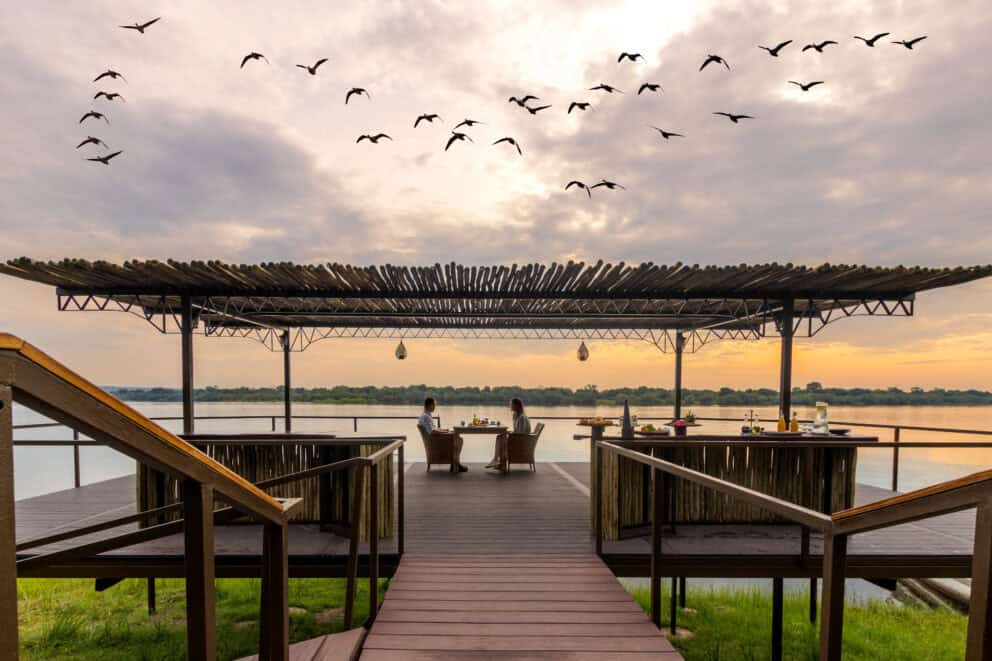 A couple having a meal on the shaded wooden deck as the sun sets over the Zambezi River at Old Drift Lodge, Zimbabwe.