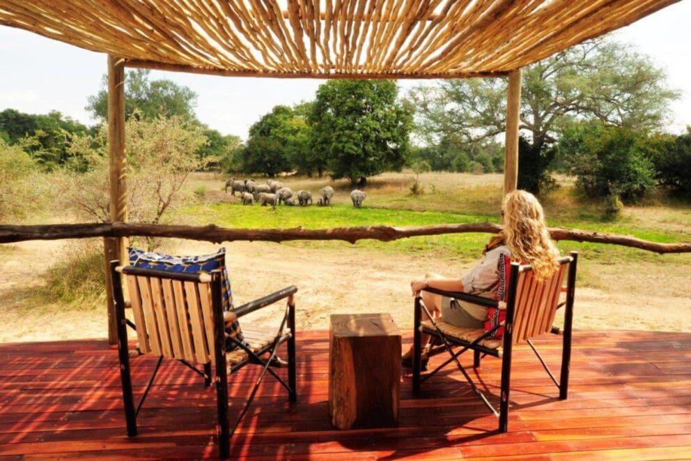 A woman sitting on a deck in a cushioned armchair watching a herd of elephants in the distance at Bilimungwe, Zambia.