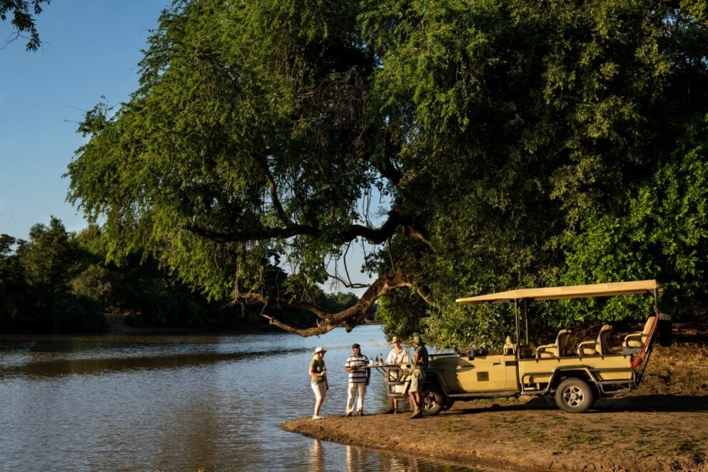 Safari travellers enjoying sundowner drinks with a safari guide along the riverbank in Mana Pools National Park at Ingwe Pan.