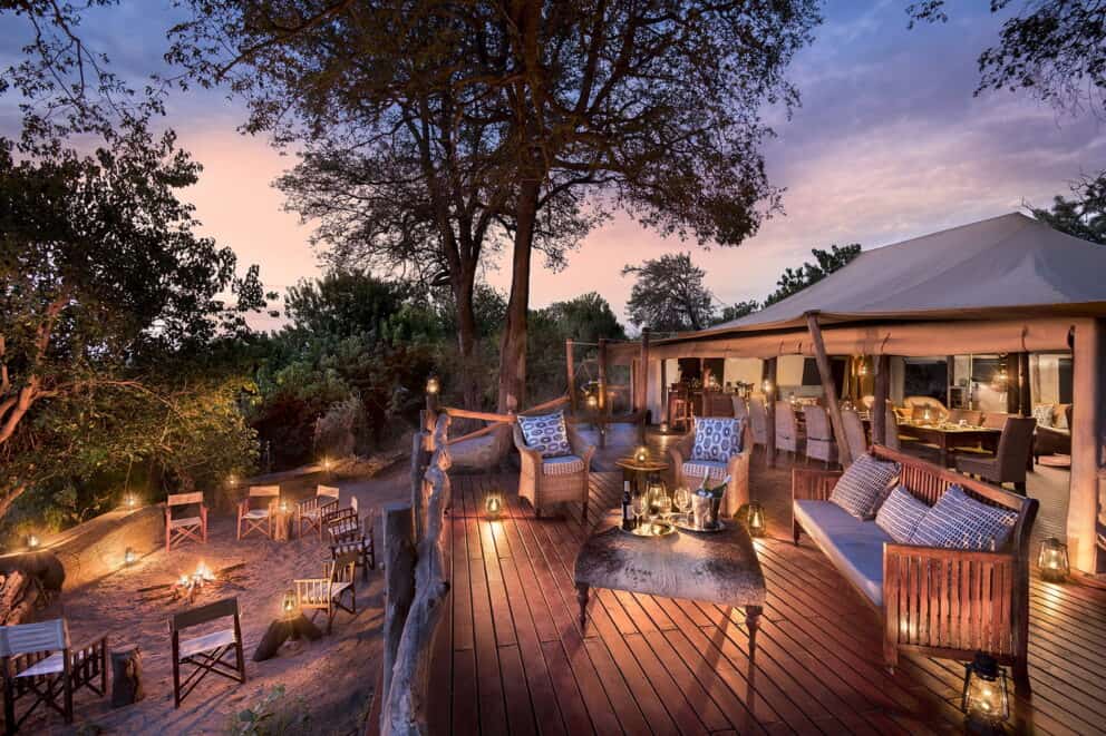 Lantern-lit elevated wooden deck with plush armchairs and a fire-lit boma below at Linyanti Bush Camp.