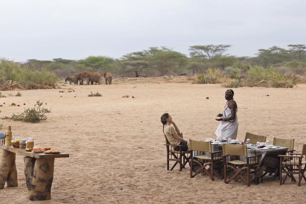 A guest seated at a breakfast table chatting to a waiter while observing elephants in the distance at Saruni Rhino, Kenya.