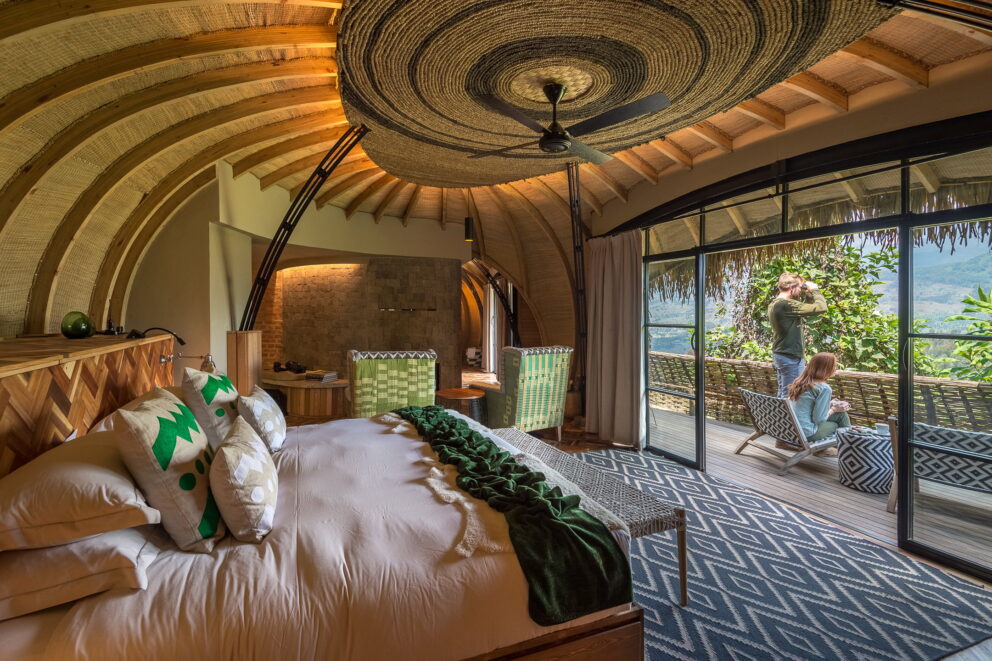 Interior view of a luxury thatched villa, opening onto a private balcony where a couple is looks out over the volcanic landscape of Volcanoes National Park landscape at Wilderness Bisate, Rwanda.