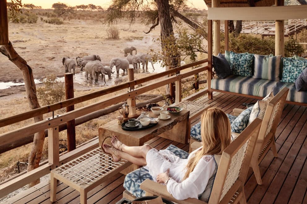 A guest relaxing in a comfy chair on an elevated wooden deck observing a herd of elephants at the waterhole at Savute Elephant Lodge, Botswana.