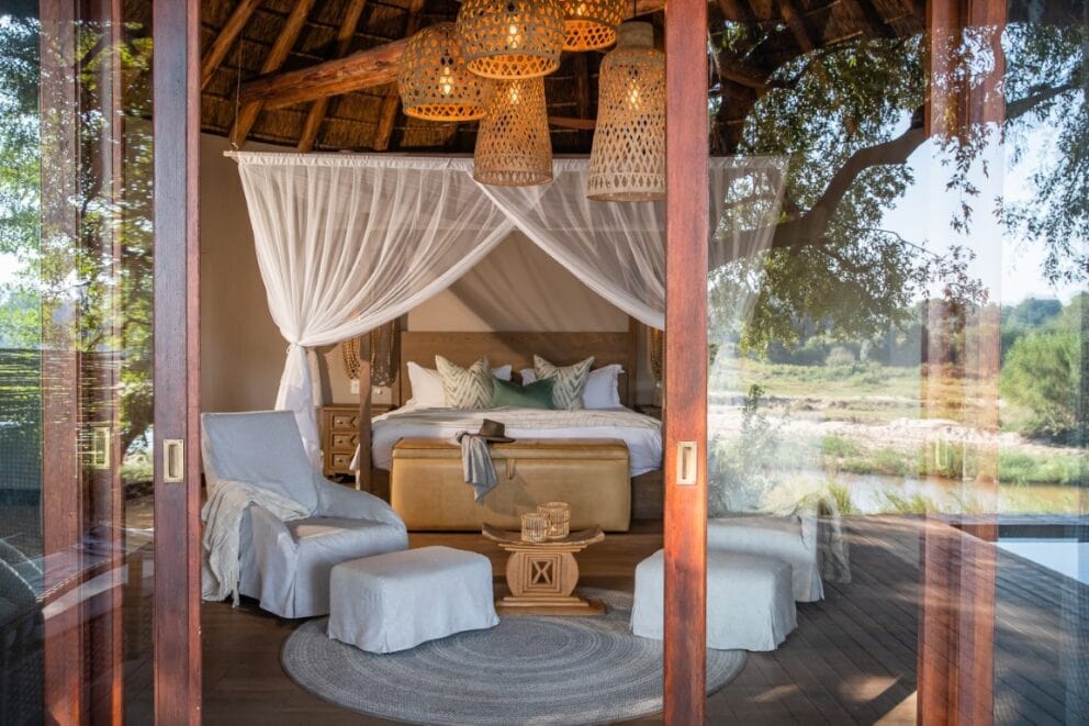 A luxury suite at Dulini River, South Africa, featuring a bed facing the outdoors and cozy armchairs under the warm glow of the hanging lamps.