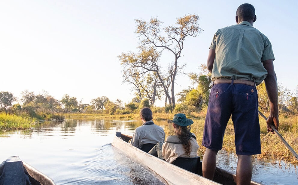 A couple sits with a guide in a mokoro as they travel down a river.