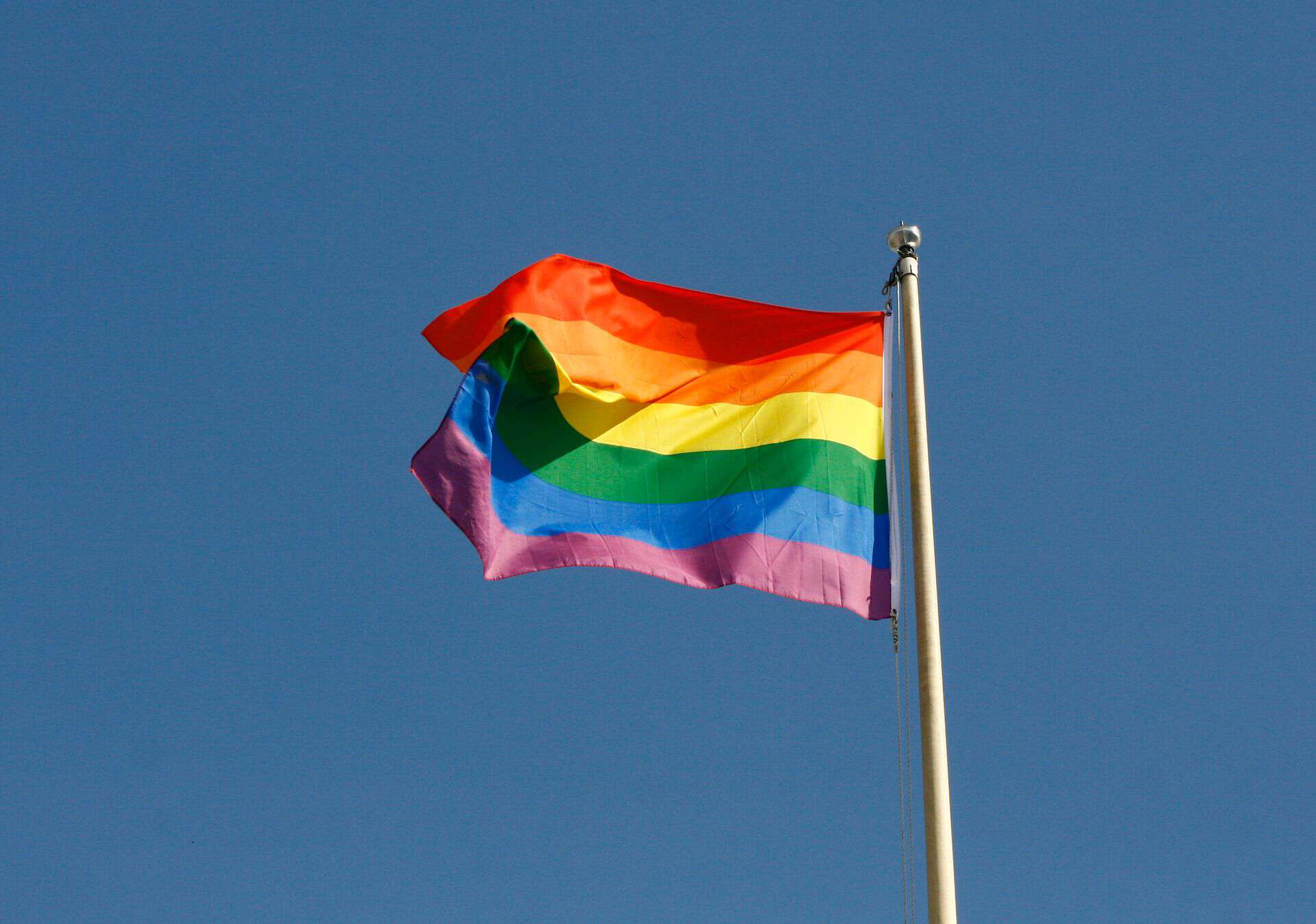 An LGBTQ Pride flag blowing in the wind on a flagpost against a clear blue sky.