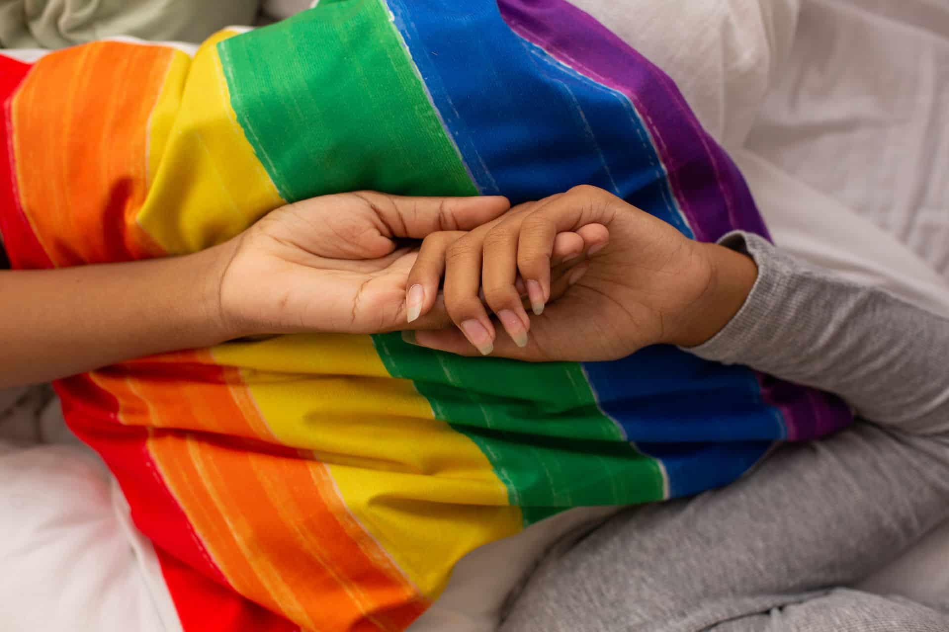 A couple holding hands, resting on top of a pillow with a LGBTQ flag pattern cover on it.