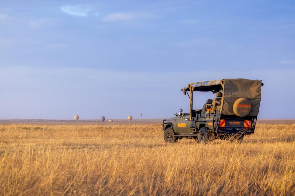 An electric safari vehicle on the open Masai Mara plain, with hot-air balloons in the distance at sunrise.