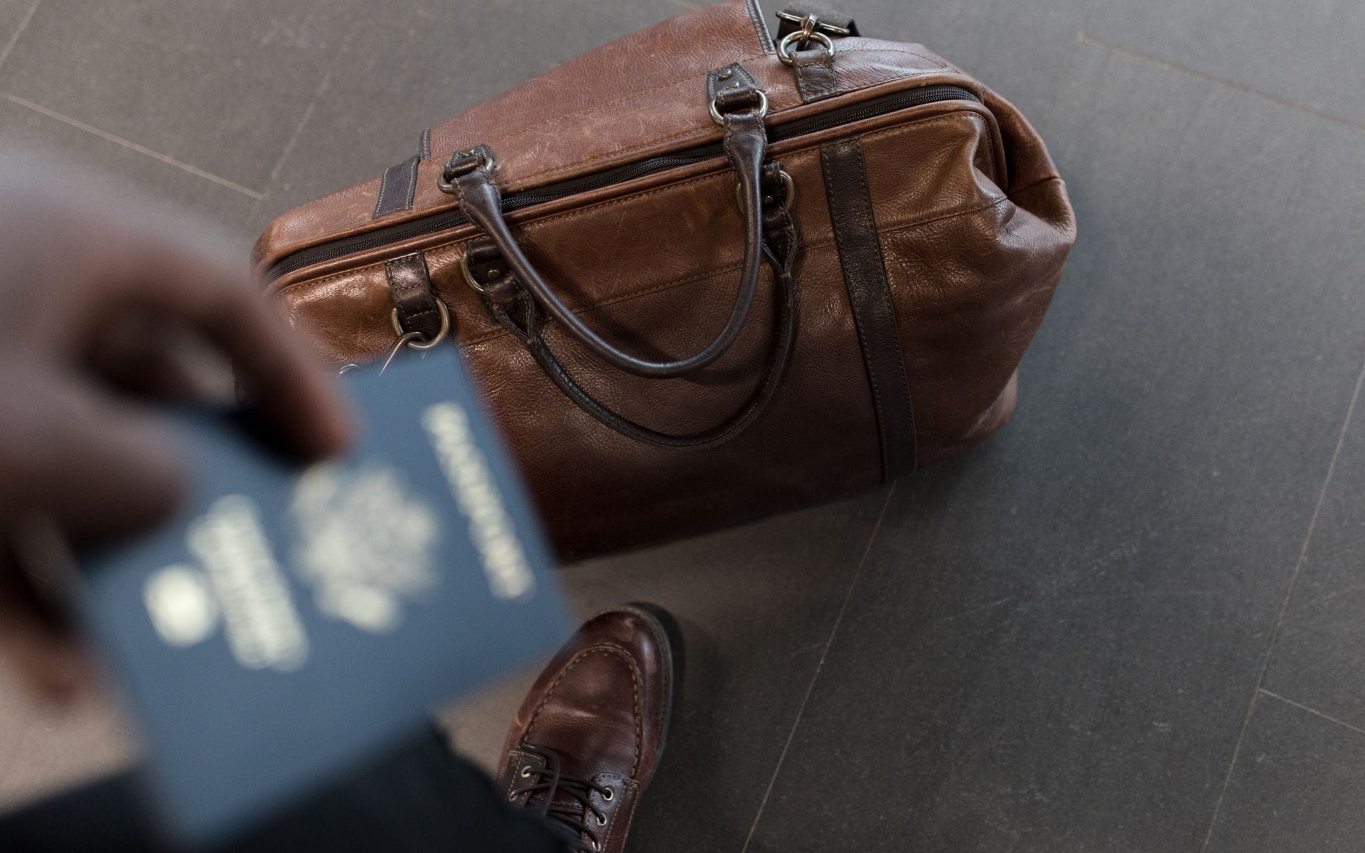 View of a brown leather duffel bag and a man holding a passport in the foreground