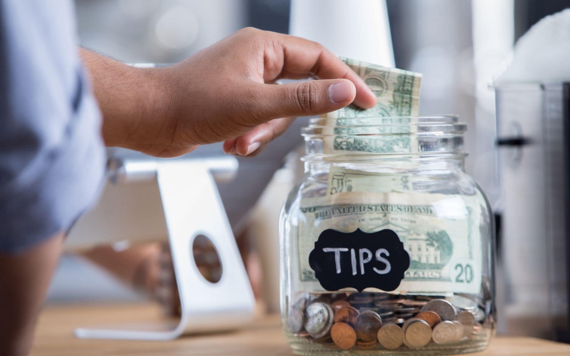 A white man's hand places a coin tip into a tipping jar.