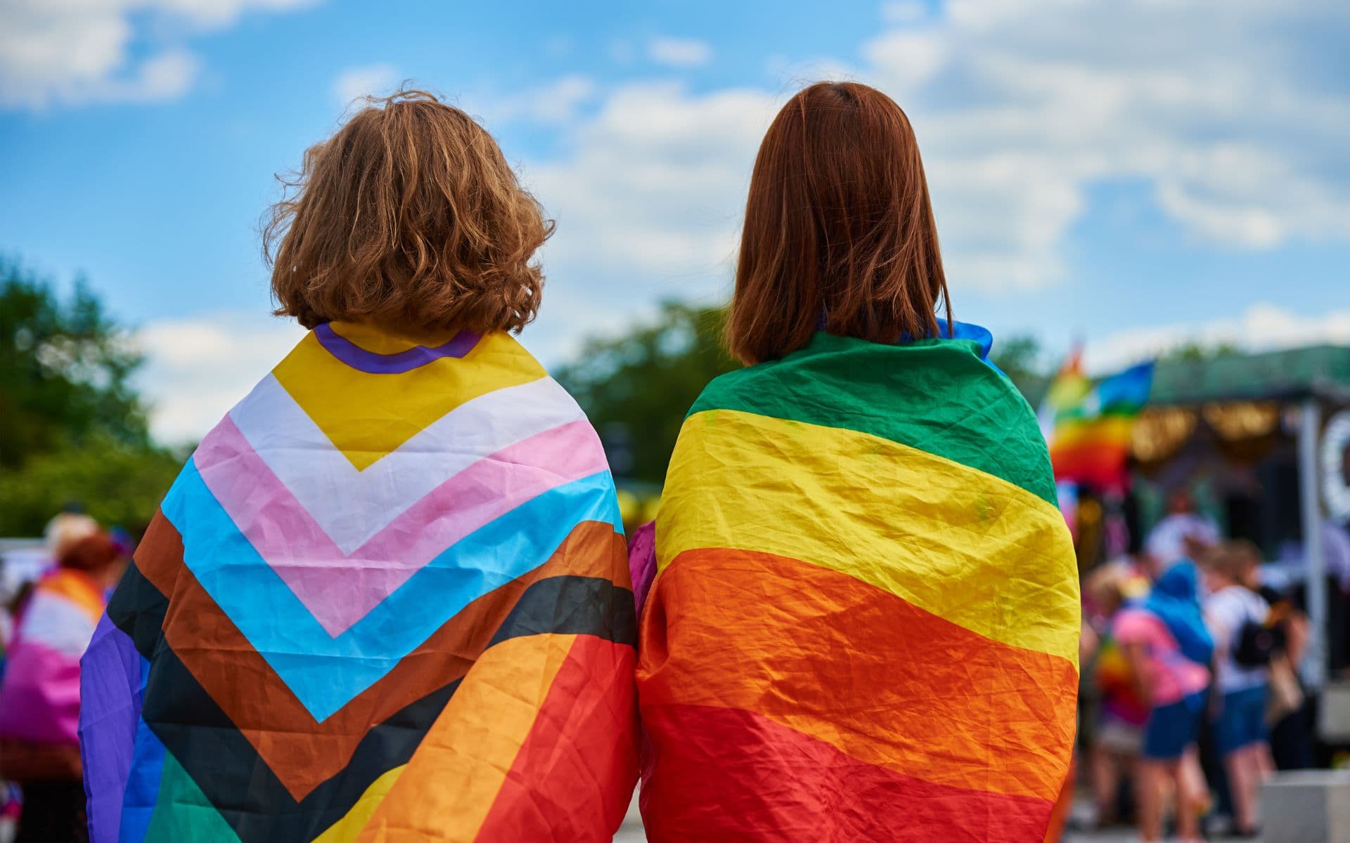 Lesbian couple at pride parade with pride flags on their backs.