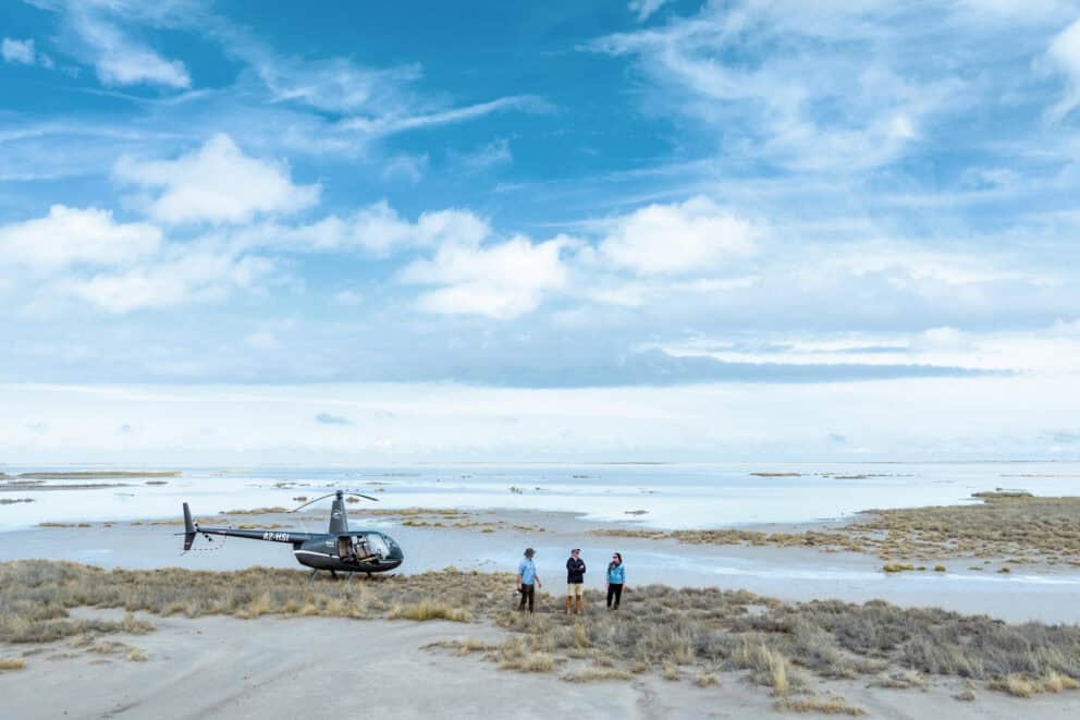 Tourists standing on a small sand island with water surrounding them and a helicopter stationed on the sand in the Okavango Delta, Botswana.
