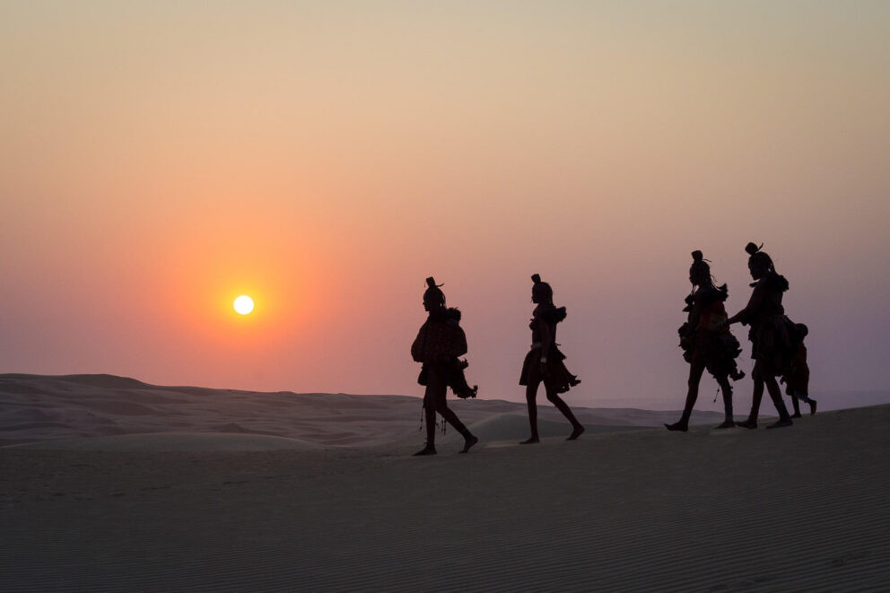 A group of Himba people walking across the desert landscape at sunset near Wilderness Serra Cafema, Namibia.